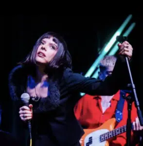 A woman with a bob haircut sings into a microphone on stage, with a musician playing a bass guitar in the background under neon lights.